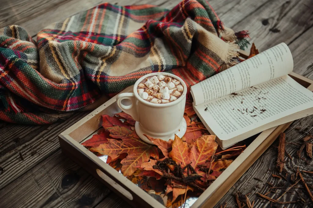 Herbstliche Decke und Blätter mit liegen bei einer gemütlichen Decke und einem Buch auf einem Holzboden.